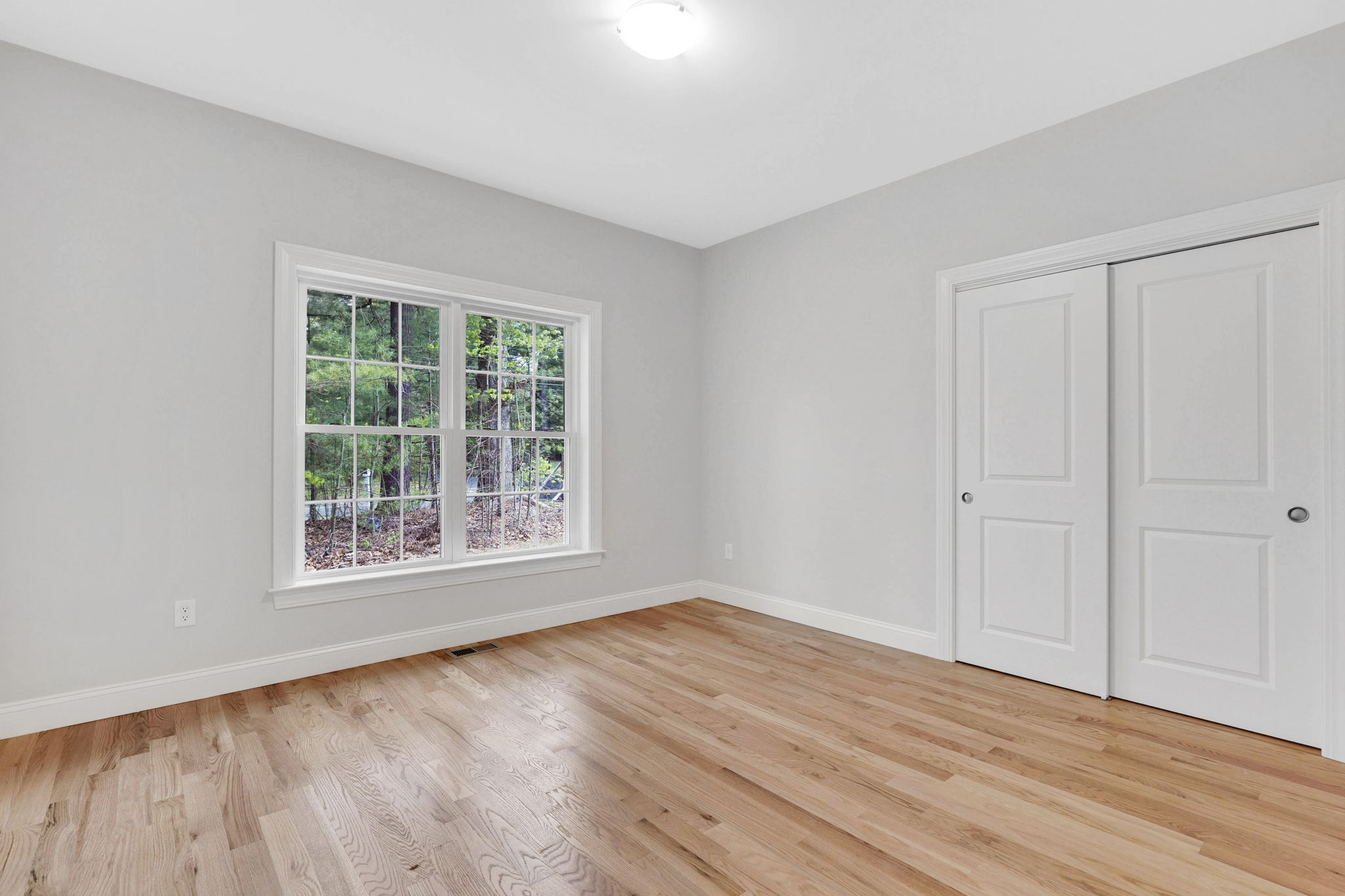 Empty bedroom with light gray walls, hardwood floors, large window and white closet doors